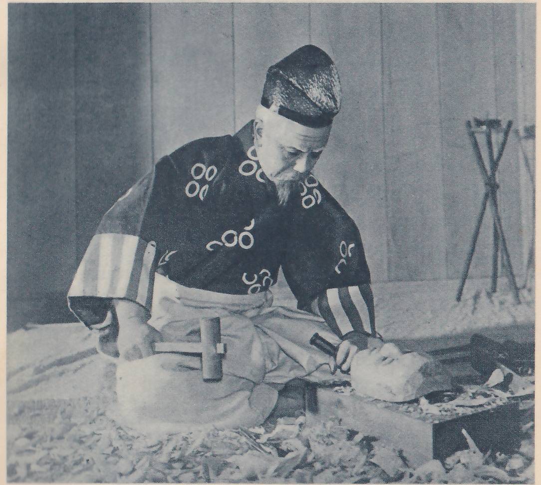 Photograph of an older man with a short gray beard and a dark cap with a band, sitting on a floor covered with pale wood shavings and looking down toward the viewer's right. He wears a dark short jacket marked with white ring-shaped patterns and sleeves with wide stripes, holds a small chisel against a pale carved face-shaped piece of wood lying on a block at the right, and has a wooden mallet on his lap. A tripod of thin poles stands at the right against a plain paneled wall.