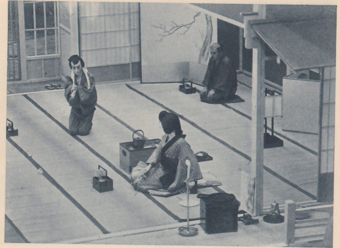 Photograph from above of a room with a mat floor and dark border strips, sliding screens at the back and a painted branch on the wall at the upper center. At the left a person with a topknot kneels with the hands clasped at the chest; at the center a person in a striped robe sits facing the left beside a low box holding a kettle; at the upper right a man in a dark robe kneels on a cushion. Small boxes with handles stand on the mats, a tall thin stand with a round foot rises at the lower center, and a roofed post and a screen stand at the right.