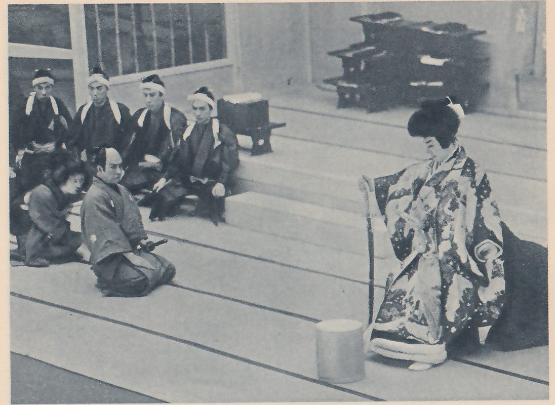 Photograph of a wide mat floor rising in steps toward the back. At the upper left a row of men in dark robes with white head bands and white cords crossing the shoulders sit in a row; in front of them a person in a dark robe bows low at the left, and beside them a man in a pale gray robe kneels facing the right. At the right a person with a high dark hair knot, in a long robe richly patterned with flowers and leaves and trimmed with a pale thick border at the hem, one hand on a long staff, and a plain round pale block stands on the floor beside them. A stack of dark low stands stands at the upper right.