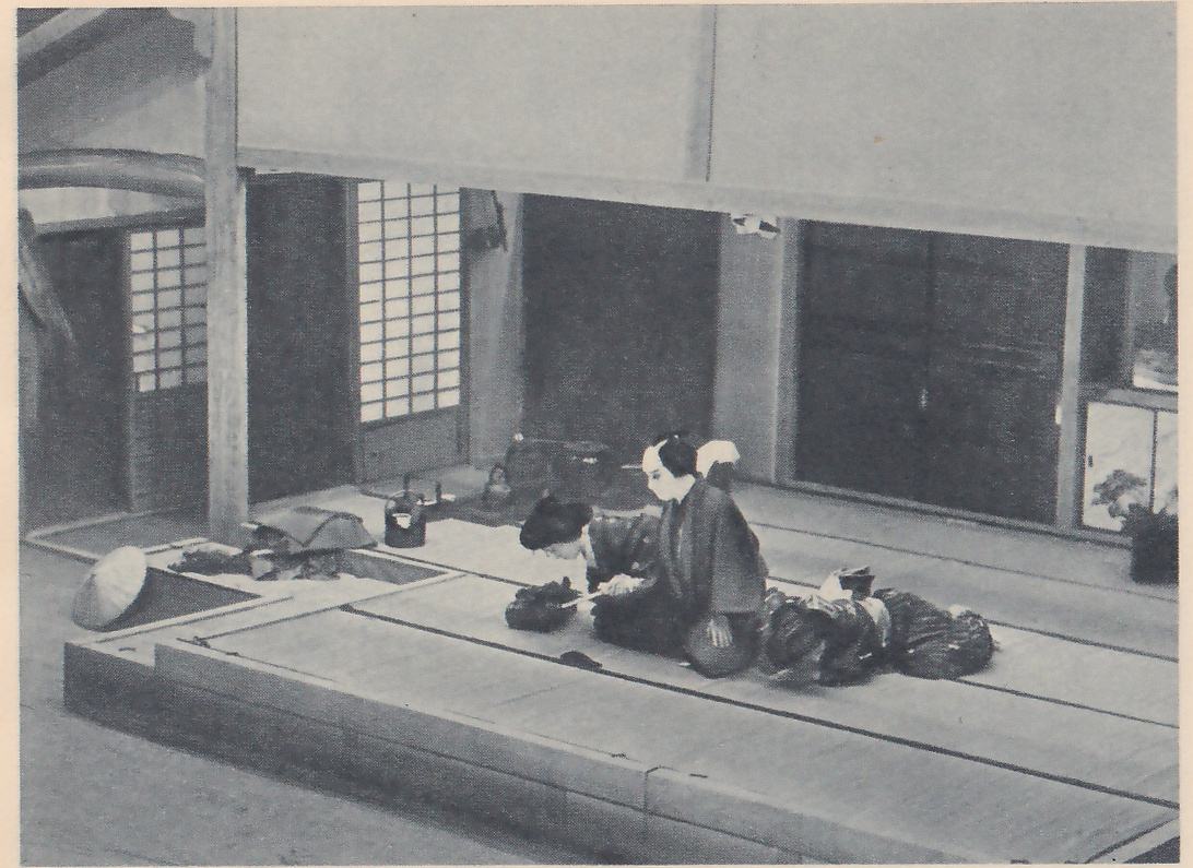 Photograph from above of a raised mat floor in a room with sliding paper screens and dark wooden doors behind. At the center two people in dark robes kneel close together, one bent far forward over a small dark bundle with the other leaning toward them; to the right a heap of dark cloth bundles lies on the mats, and at the left a square recess in the floor, a kettle and a wide conical hat lie near the edge. A potted plant stands at the right edge, and a large pale panel hangs across the top.
