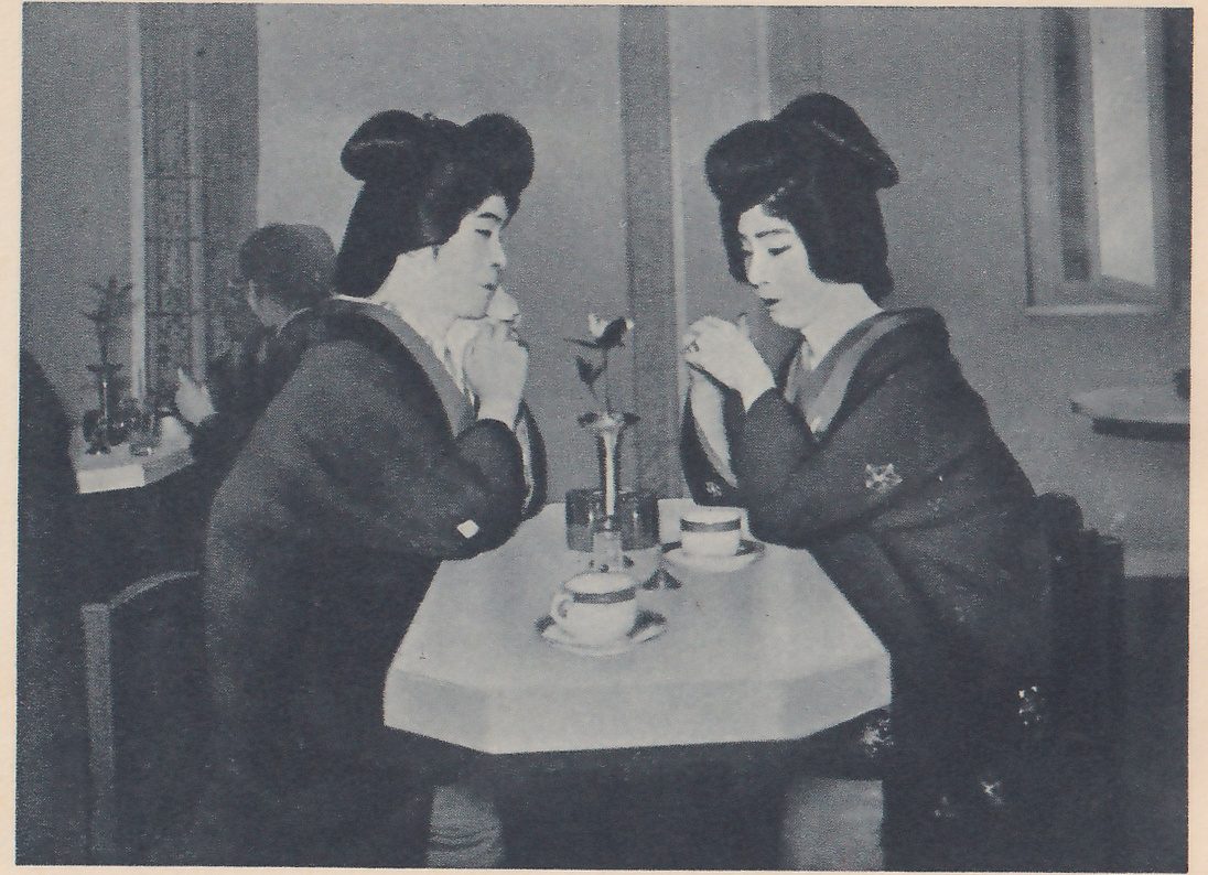 Photograph of two people with large, dark, upswept hair sitting at either side of a pale table with cut corners, leaning toward each other with their hands raised near their chins. Both wear dark robes with small patterns. On the table stand two cups on saucers and a tall slim vase holding a single bloom. Behind them at the left another person sits at a small table beside a window, and a tall window shows at the upper right.