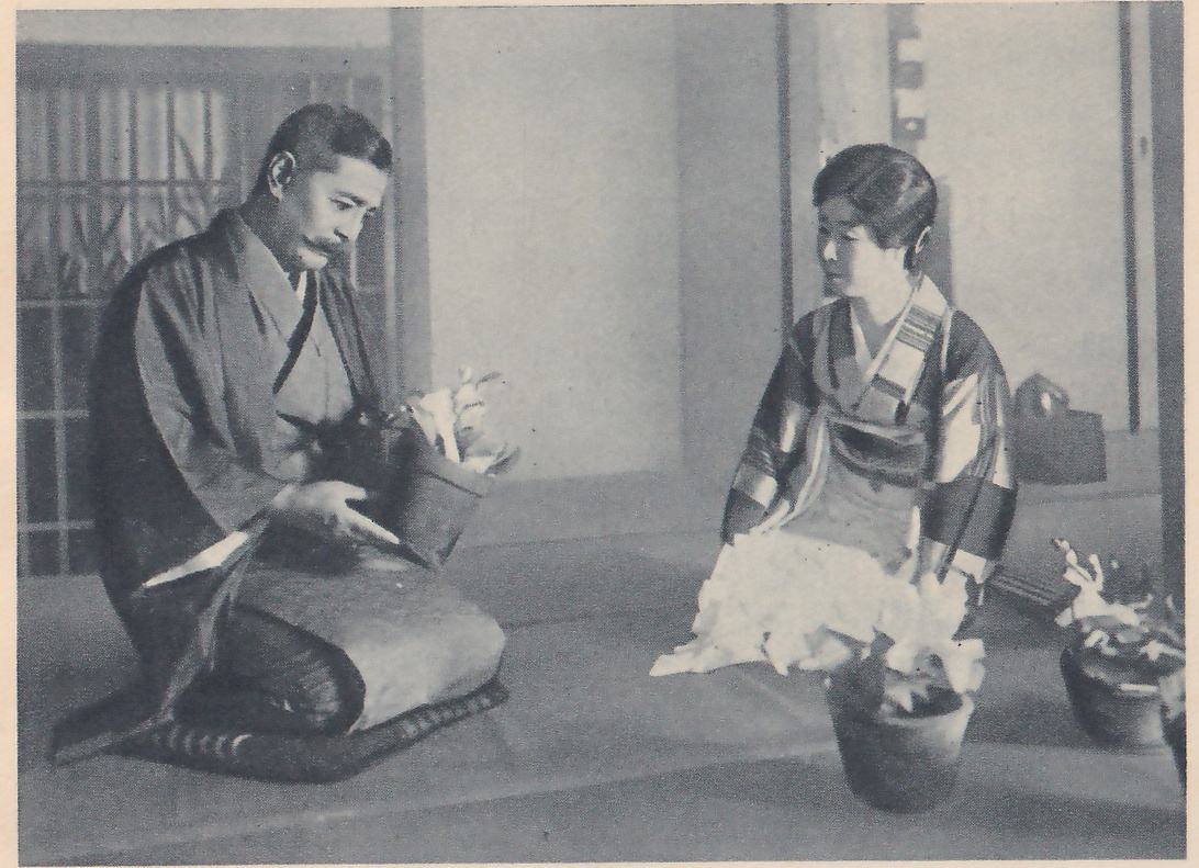 Photograph of two people kneeling on a mat floor facing each other. At the left a man with a moustache and short dark hair sits on a striped cushion in a dark kimono, holding a dark pot with white flowers in both hands and looking down at it. At the right a woman in a kimono with wide bands of pattern sits looking down toward a large pot of white-flowered plants on the floor in front of her; more pots stand at the lower right. Behind them are plain walls with sliding screens and a window with a lattice at the upper left.