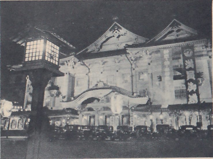 Night photograph of a large building front lit from below: two steep gabled roofs with ornamental ends, a curved canopy over the entrance at the center, and a tall narrow banner decorated with flower shapes at the right. A square lantern on a thick post, its panes glowing, stands in the left foreground, and a row of parked cars lines the street in front of the entrance.