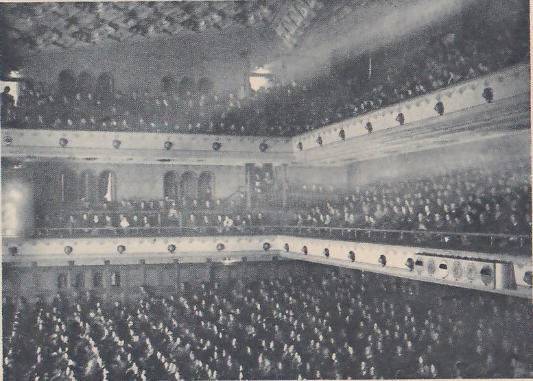 Photograph of a large hall seen from one side, with the floor and two balcony levels packed with rows of seated people. White balcony fronts studded with small round lights run along the near and far walls, arched openings show along the back wall, a patterned ceiling shows at the upper left, and a haze of light falls from the upper right.