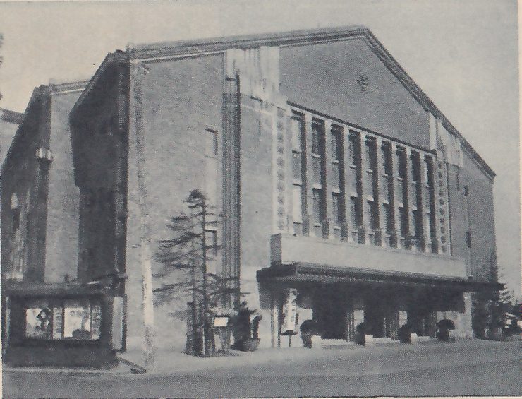 Photograph of the front of a large building with a tall gabled front and a stepped wing at the left. A band of tall narrow windows in a pale frame runs across the upper front above a flat canopy over the entrance, with vertical decorative strips down the wall. A slender tree and small potted trees stand on the pavement in front, and a small kiosk with posters stands at the lower left.