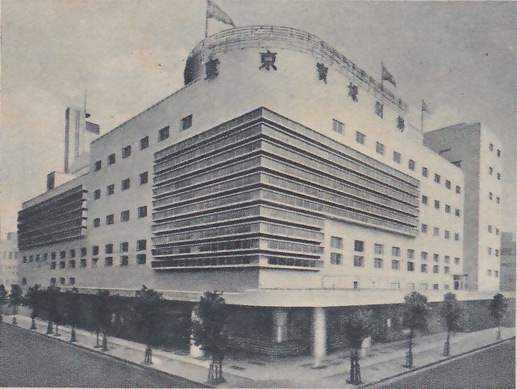Photograph of a large pale modern building seen from a street corner, with a rounded top bearing the theatre's name in a row of large characters, 東京寶塚劇場, and flags on poles, wide horizontal ribbed bands across the front, rows of small square windows, and a roofed ground floor on round columns. A row of young trees stands along the pavement in front, and a taller block rises at the right.