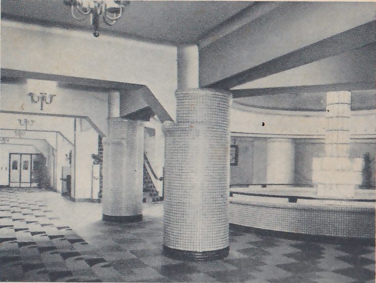 Photograph of a wide lobby with two round columns faced with small square tiles in the foreground and a checkered floor. A chandelier hangs at the upper left, a corridor with wall lamps and a pair of doors lies at the left, and at the right a curved low wall surrounds a tall stepped block of pale squares. A beam crosses the ceiling at the upper right.