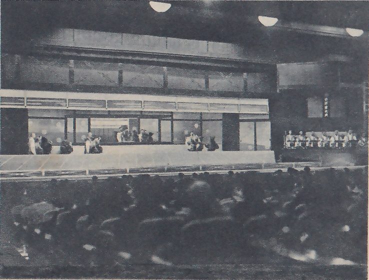 Dark photograph taken from among a crowd of blurred heads in the foreground toward a wide, lit, low raised floor backed by long window-like openings, with small figures standing and sitting at the left and center of it. At the right a row of people in light clothes sits along a raised ledge below a small upright sign, and round lights show at the top of the ceiling.