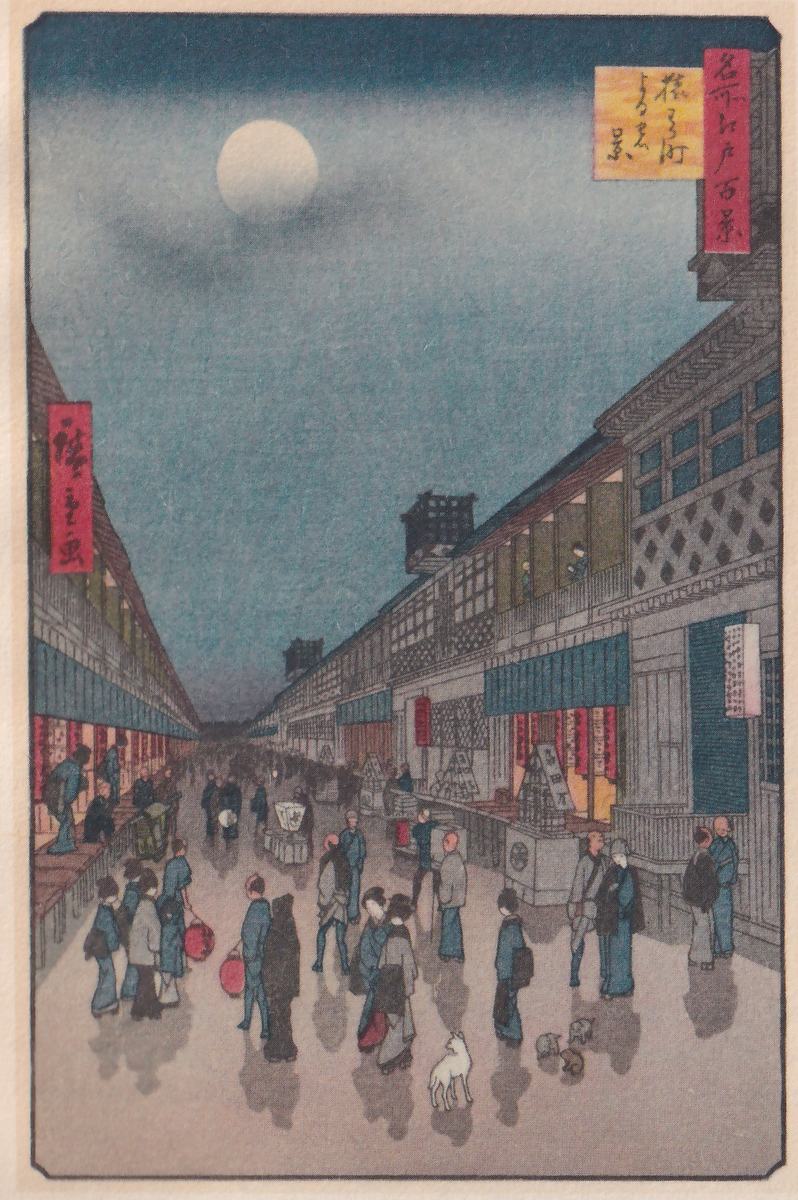 Colored print of a long street at night, seen straight down its length between two rows of buildings with latticed fronts and blue awnings, receding to a faint point under a dark blue sky with a pale round moon at the upper left and streaks of cloud. People in blue and gray robes walk along the street, a few carrying red round objects, with a white dog at the lower center and smaller animals at the lower right. At the upper right a red cartouche reads 名所江戸百景, the series One Hundred Famous Views of Edo, beside a yellow card with the print's title, 猿わか町よるの景, Saruwaka-machi by night; a red cartouche at the left carries the signature, 廣重画, Hiroshige.