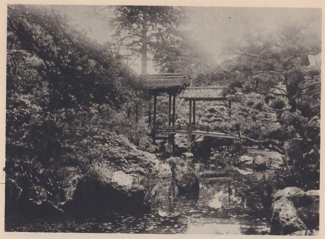 Photograph of a pond in a misty, steep-sided garden, with a roofed wooden bridge with two pitched roofs crossing it at the center, a rocky wooded bank at the left, large rocks at the water’s edge at the lower right, and pines at the upper right.