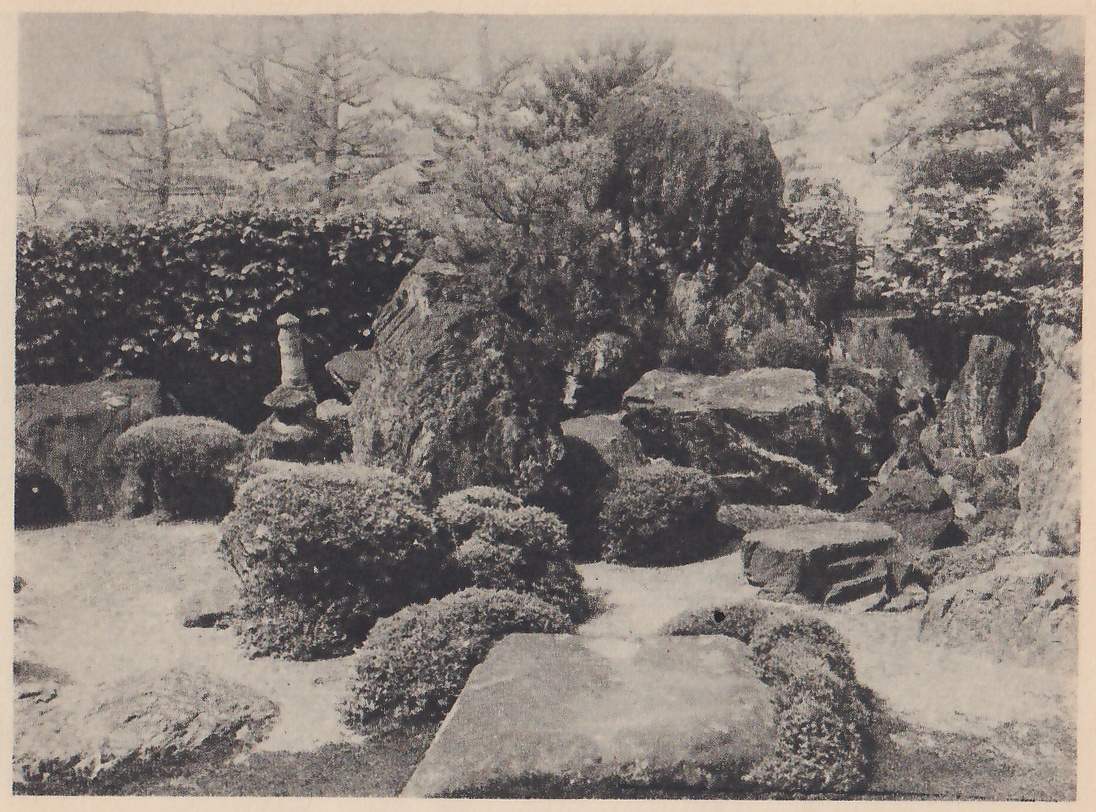 Photograph of a rock garden: tall rough rocks and flat slabs piled at the back, rounded clipped shrubs in front of them, a small stone pagoda or lantern at the left against a clipped hedge, and a large flat slab lying on the pale ground in the foreground with pines and trees at the top.