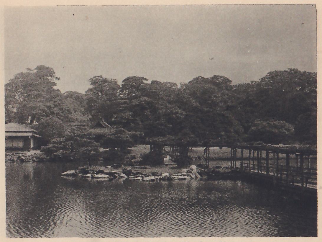 Photograph across a rippled pond to a low rocky island with a roofed building and pines behind it, a long wooden bridge on posts with railings running across the right, and a small house with a tiled roof at the far left among dark pines.