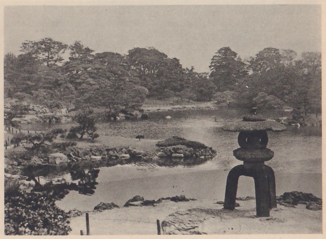 Photograph over a pond garden with a tall stone lantern in the right foreground, its wide round cap on a rounded body above stout curved legs, standing on a bare sandy bank; beyond it a pond with low rocky islets and a pine, and wooded shores under a pale sky.