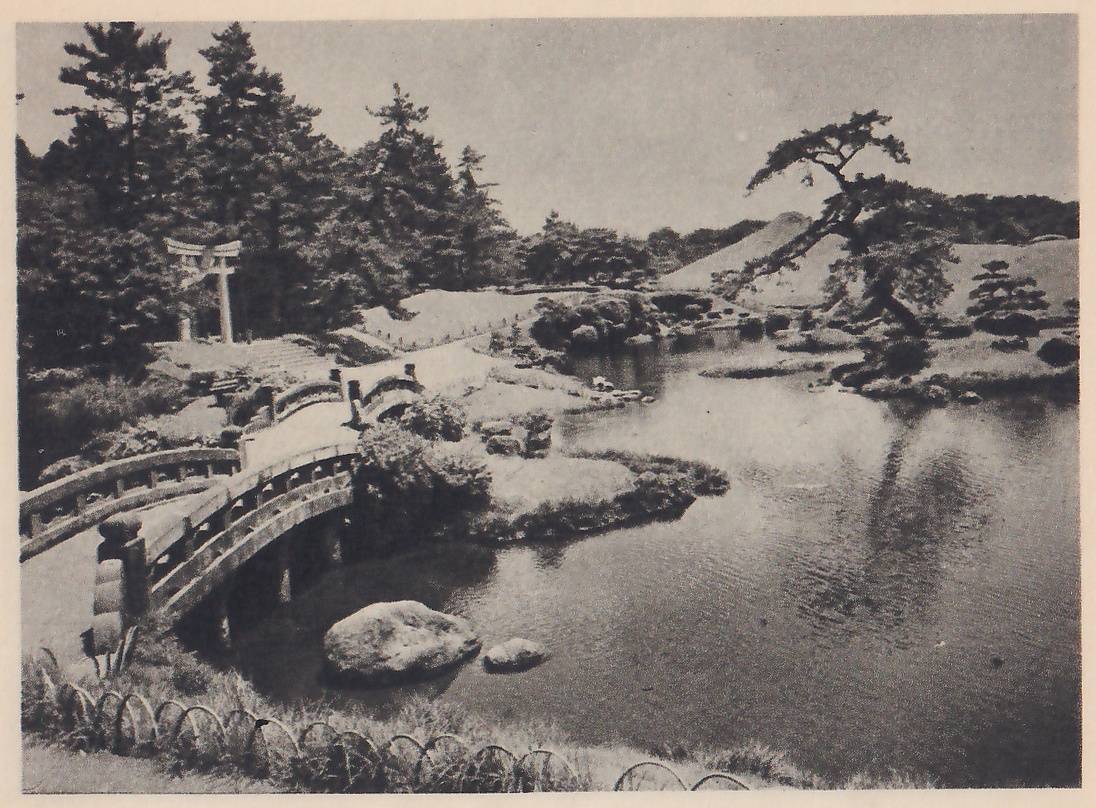 Photograph over a wide pond from a bank at the lower left: a steep arched wooden bridge with a railing and a second smaller arched bridge lead across an inlet toward a small torii gate among tall pines at the upper left; a large rounded rock stands in the water below the bridge, and a leaning pine and a smooth grassy mound rise on the far shore at the right.