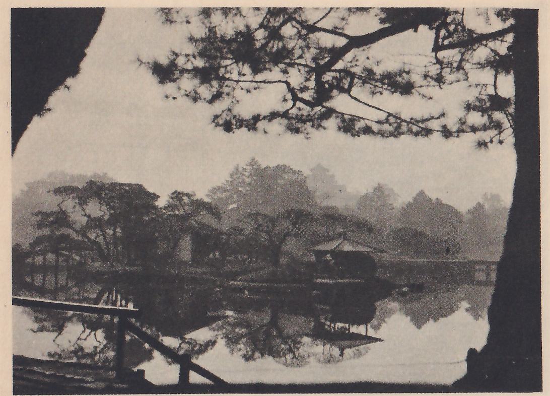 Photograph in misty light through the dark trunk and branches of a pine at the right and upper left: across a still pond lie a small island of pines with a pavilion with a pointed roof at the right, a low bridge at the left, and pale trees in haze; a rail of poles crosses the left foreground.