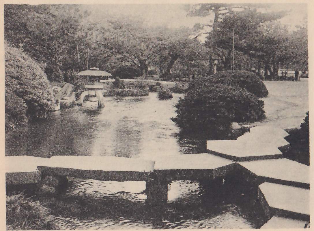 Photograph of a stone slab bridge of several flat slabs crossing a rippled stream in the foreground, with a zigzag of six-sided stepping-slabs leading off at the right; beyond it a pond with a stone lantern with a wide cap beside rocks at the left, a rounded clipped shrub at the right, and pines and other trees behind, with a few people far off at the upper right.