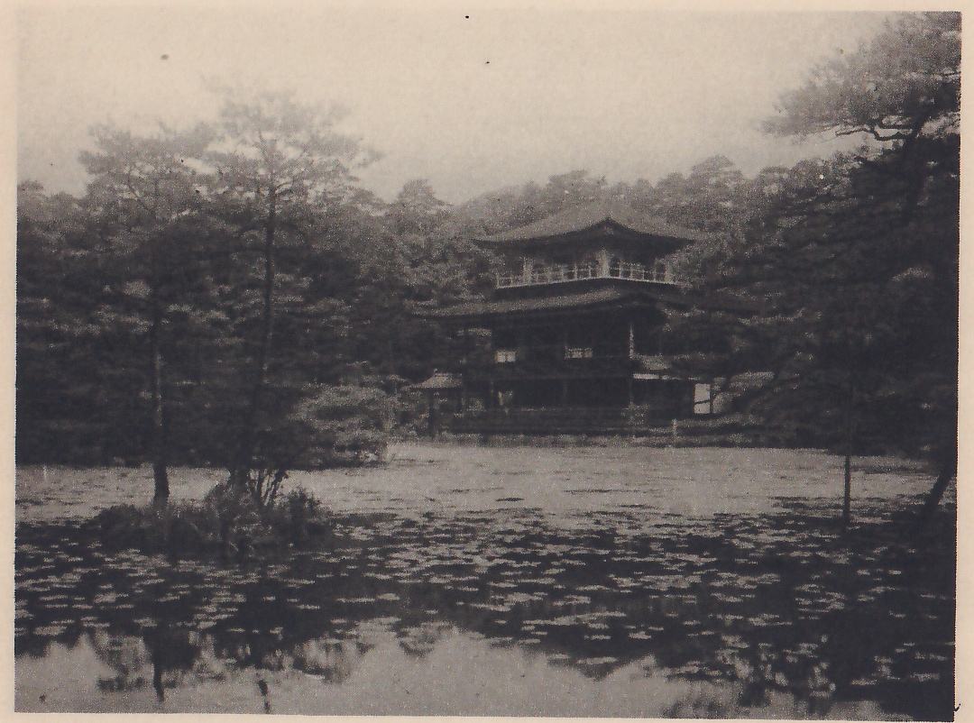 Photograph across a pond covered with floating leaves to a three-story pavilion with curved, tiered roofs and a railed upper floor, among pine trees; a small island with a slender pine stands in the water at the left.