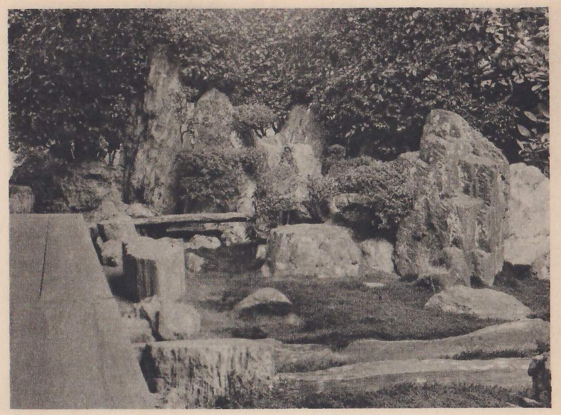 Photograph of a garden of rocks: tall upright stones and low rounded boulders stand among clipped shrubs and moss, with a flat slab laid across a gap in the middle, a dark hedge behind, and the edge of a wooden floor at the left.