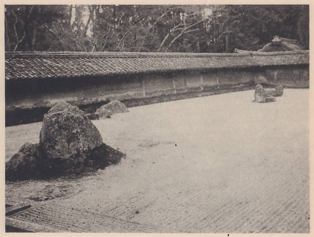 Photograph of a wide stretch of pale sand marked with fine lines, enclosed by a long low wall with a tiled top, with trees rising behind it. A group of rocks stands at the left, and a smaller group of stones lies near the wall at the right.