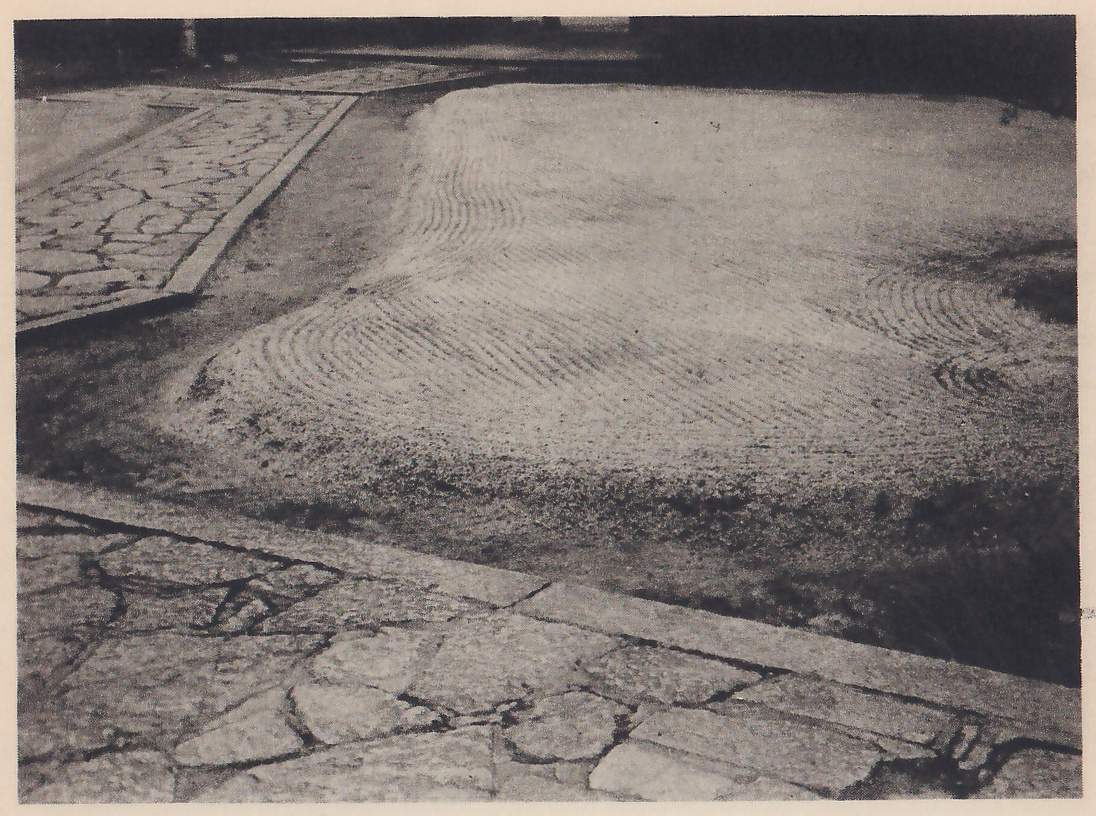 Photograph looking down at a bed of pale sand raked into curving lines, bordered along the left and across the foreground by paths of flat, irregular paving stones with raised stone edges.