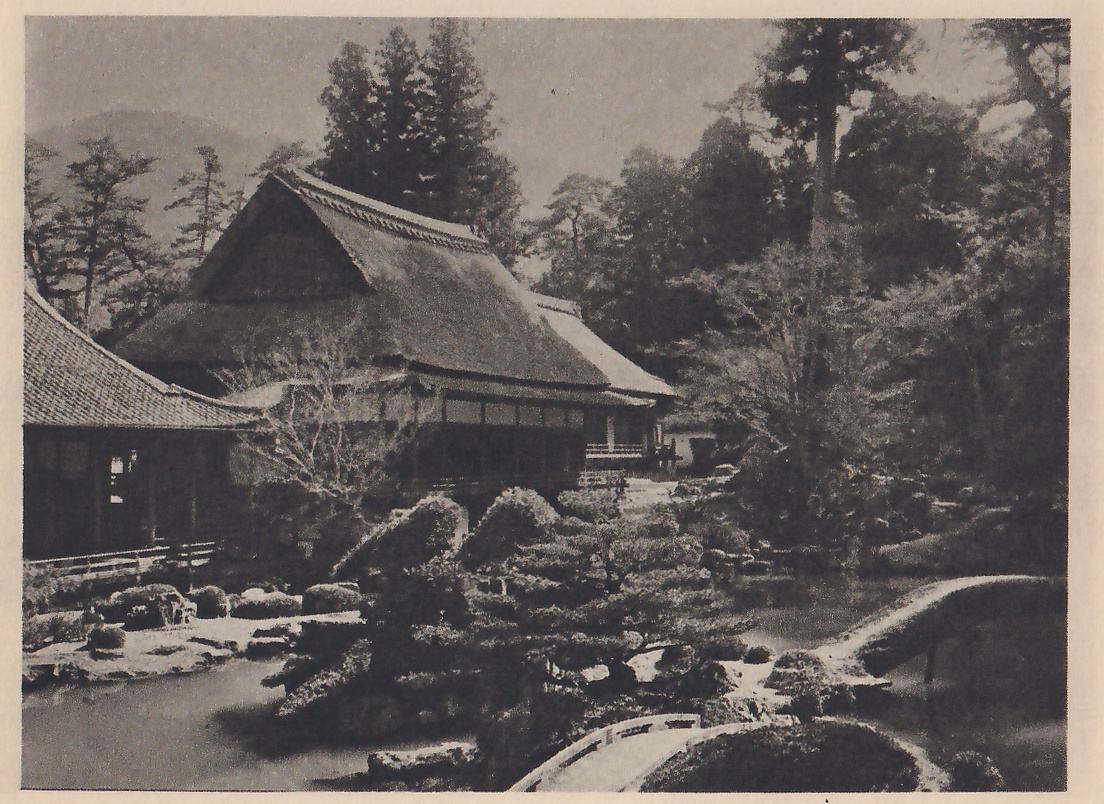 Photograph of a pond garden in front of a large thatched-roof house with a tiled-roof building at the left; clipped pines and rounded shrubs stand at the water’s edge, an arched bridge with a railing crosses the water in the foreground, a second low arched bridge shows at the right, and tall trees and a mountain rise behind.