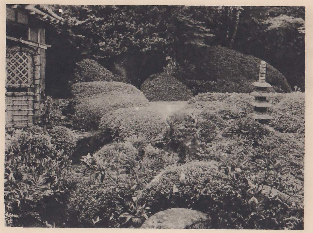 Photograph of rounded, clipped shrubs in banks across a garden, with a rough rock among them at the center and a tall stone tower of several stacked tiers at the right. The corner of a building with a lattice window and a bamboo-bound lower part stands at the left, and trees rise behind.