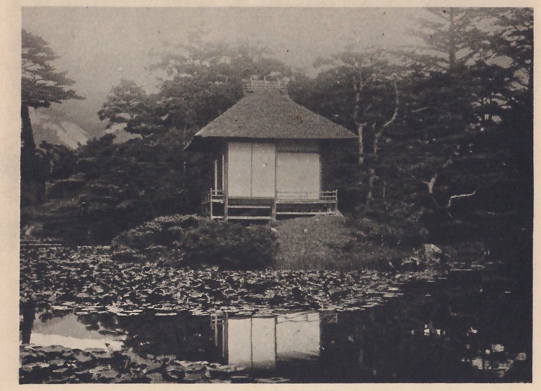 Photograph of a small thatched-roof tea-house with sliding paper doors and a railed verandah, standing on a low grassy bank above a pond covered with floating leaves; pines and other trees surround it, and the house is reflected in the water.
