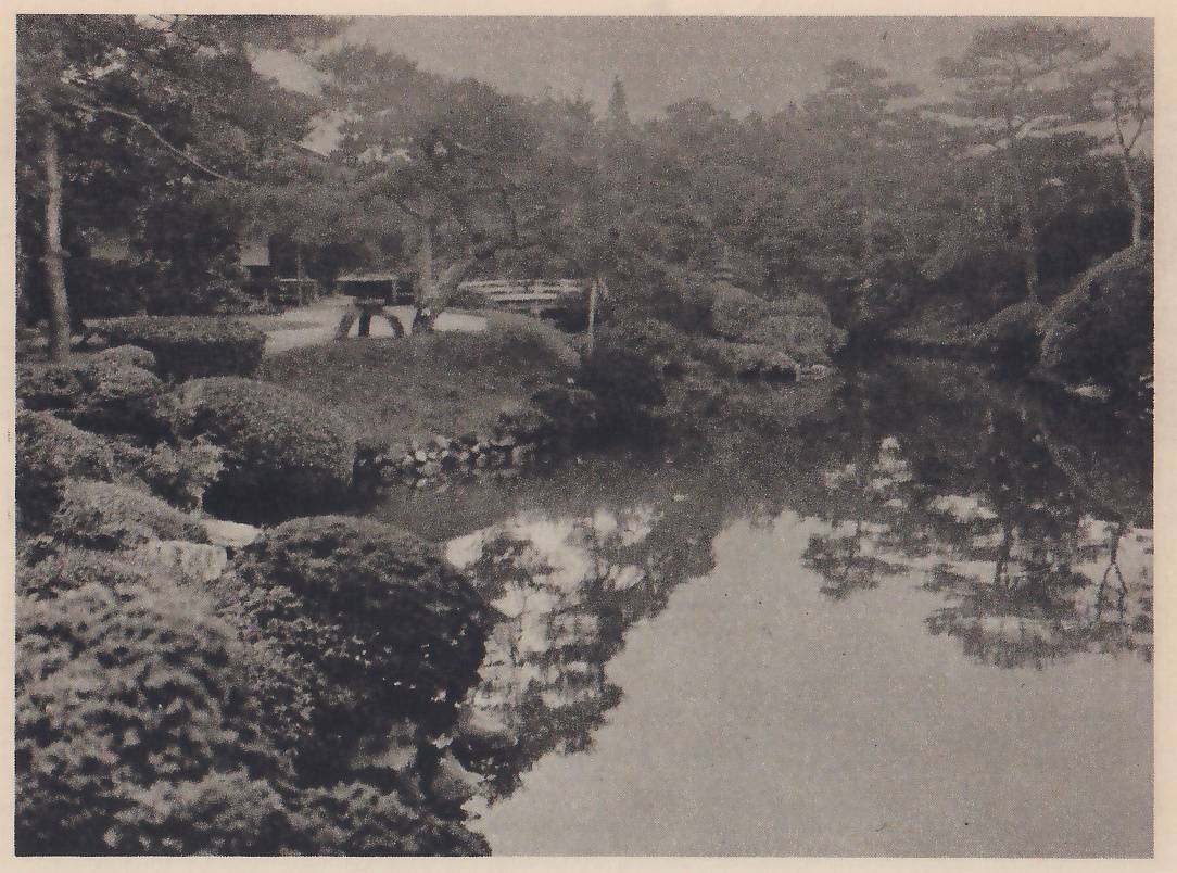 Photograph across a still pond with pines reflected in it, to a far bank with a low wooden bridge, a pale open area and the roof of a building among trees; rounded clipped shrubs fill the near bank at the left, with a low stone edging beyond them.