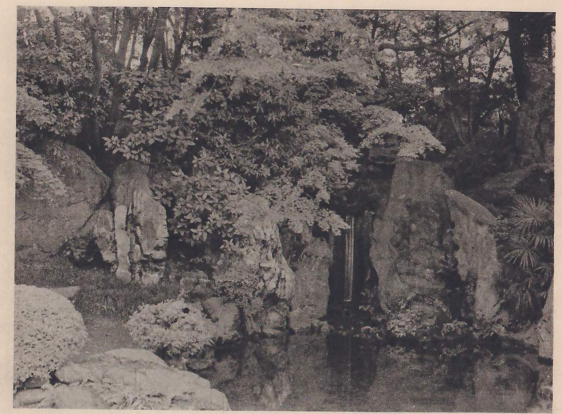 Photograph of tall upright rocks standing among shrubs and trees at the edge of a pond, with a narrow waterfall dropping between two of the rocks at the center into the dark water; a rounded clipped shrub and a flat rock lie in the lower left foreground.