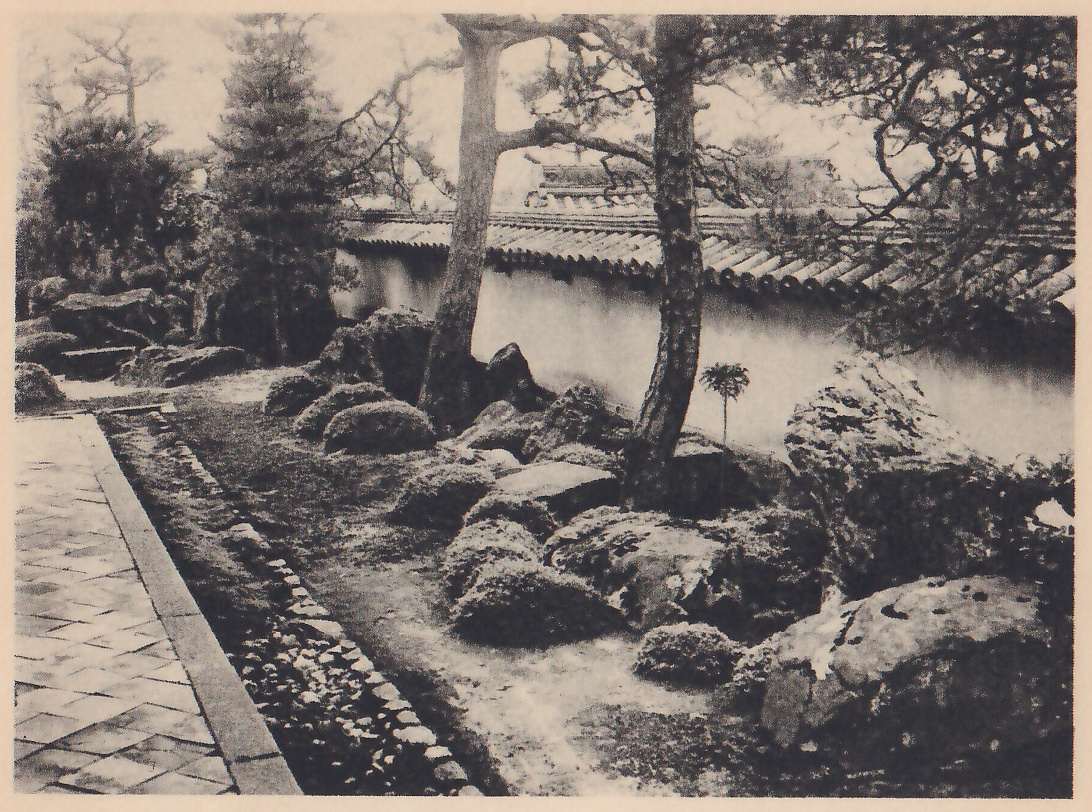 Photograph of a narrow garden bed of large rocks and rounded clipped shrubs along a long white wall with a tiled top, with two pines standing in front of the wall. A paved walk lies at the left, separated from the bed by a stone edging and a strip of small stones.