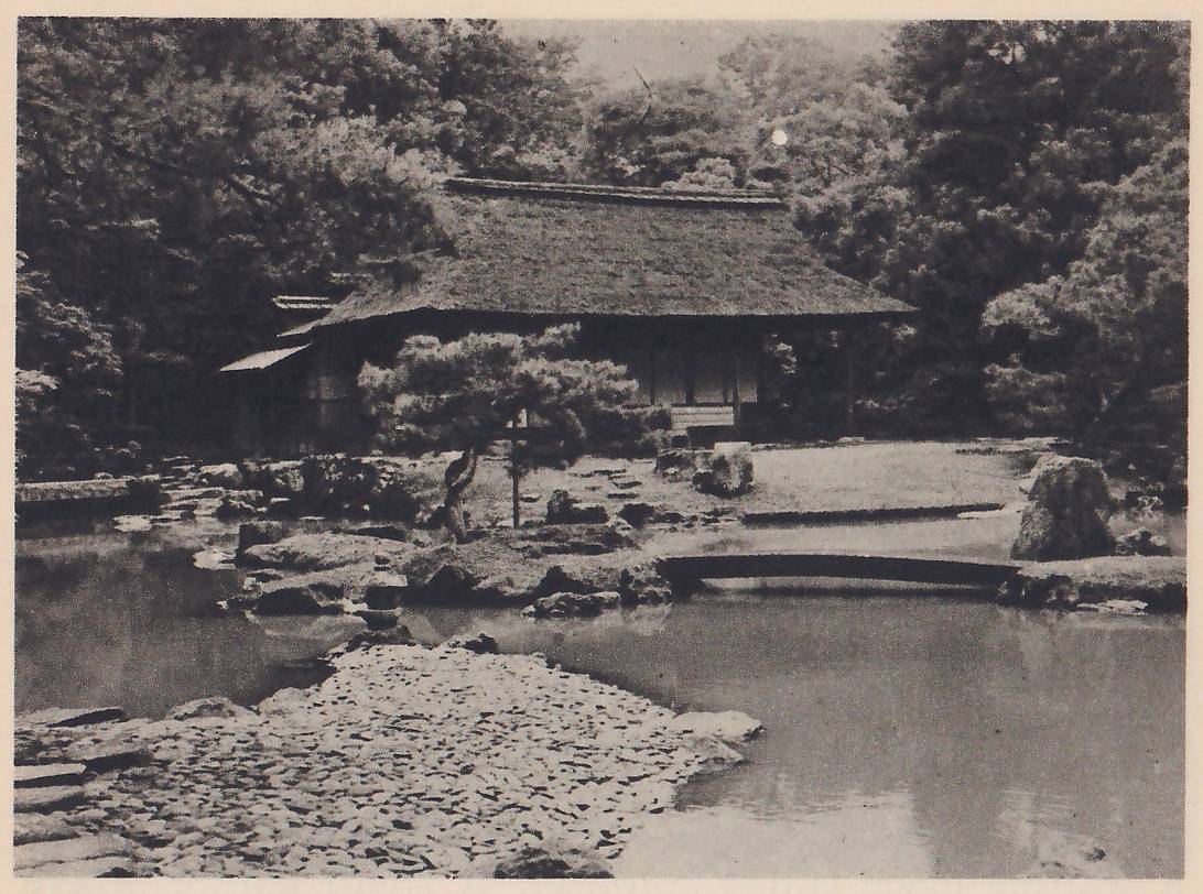 Photograph across a pond to a thatched-roof house among trees on the far bank, with a pine and rocks in front of it and a low arched bridge crossing the water at the right. A spit of small flat stones fills the lower left foreground.