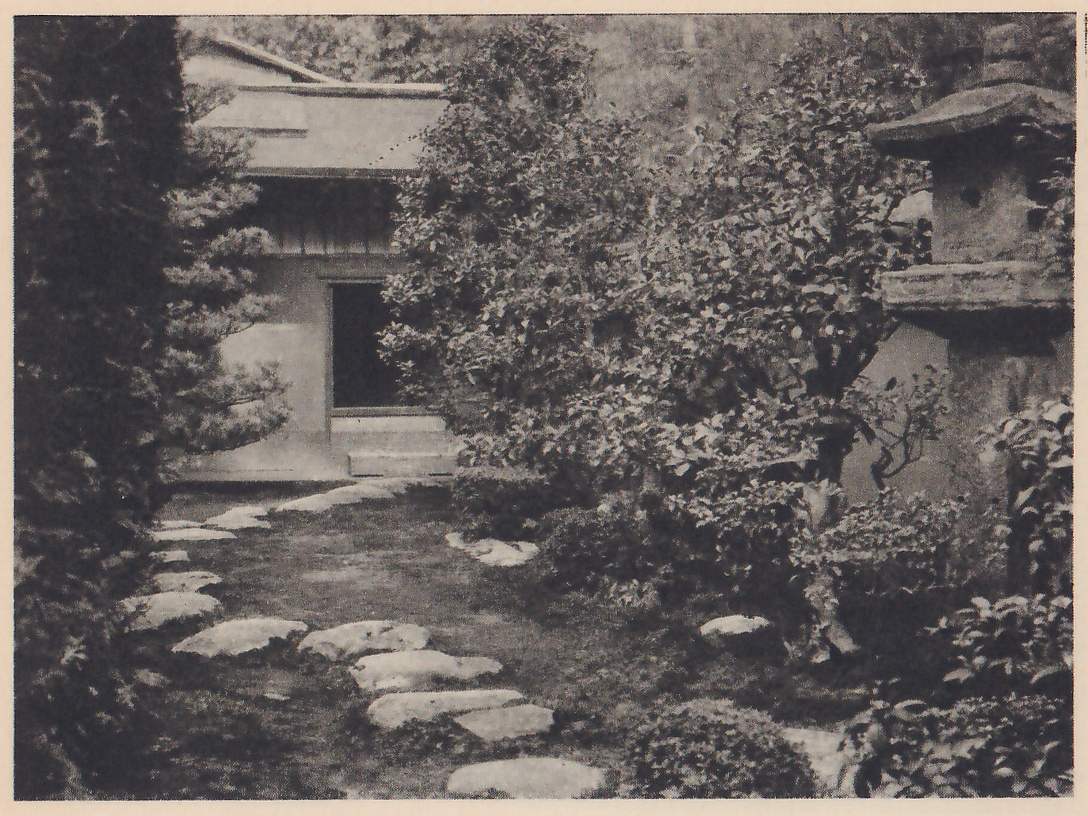 Photograph of a path of irregular stepping-stones set in moss, leading back to a plain building with a low, dark doorway; shrubs and a clipped evergreen stand on either side and a stone lantern with a thick cap stands at the right edge.