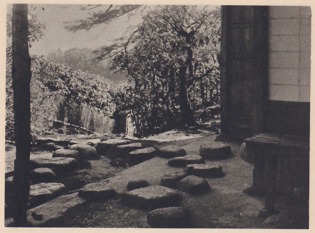 Photograph of a garden path of large, rounded stepping-stones set in moss, passing between a thin tree trunk at the left foreground and the wall of a building with paper doors at the right; a hedge and trees rise along the far side, and a hazy hill shows behind.