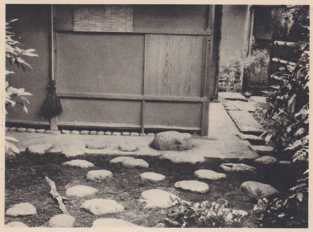 Photograph of a plain wall of the tea-room with a wooden panel door and a bundle of dark fiber or hair hanging at its left edge, above a row of small stones set along its foot. Rounded stepping-stones cross the mossy ground in front, a larger stone sits by the wall, and paving leads off at the right between shrubs.