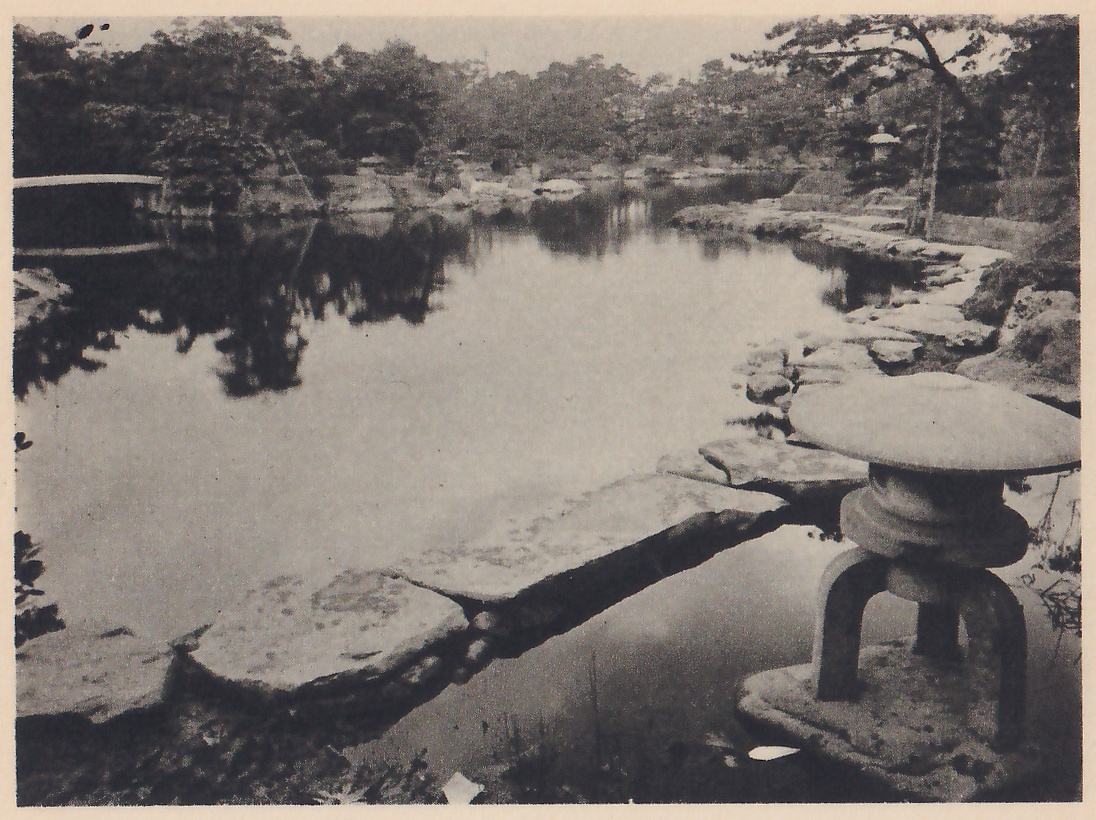 Photograph of a large still pond with trees and a low bridge reflected in it, seen along a shore of flat slabs of rock that run out into the water at the lower left. A stone lantern with a wide round cap and curved legs stands on a rock at the lower right, and another lantern shows among trees on the far shore at the upper right.