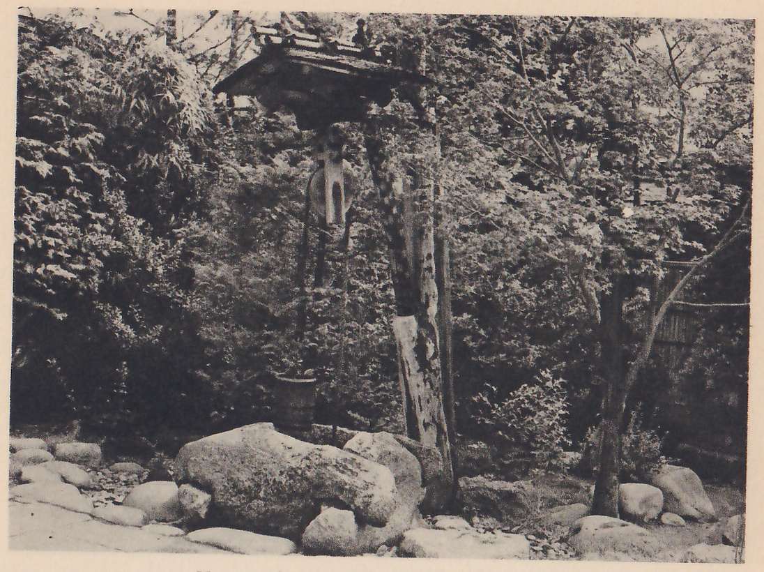 Photograph of a well frame in a wooded garden: a tall weathered post carries a small tiled roof with a pulley wheel hung beneath it, and a wooden bucket stands on the ground at the left of the post. A large rounded rock and smaller stones lie at its foot, with shrubs and trees all around and a bamboo fence at the right.