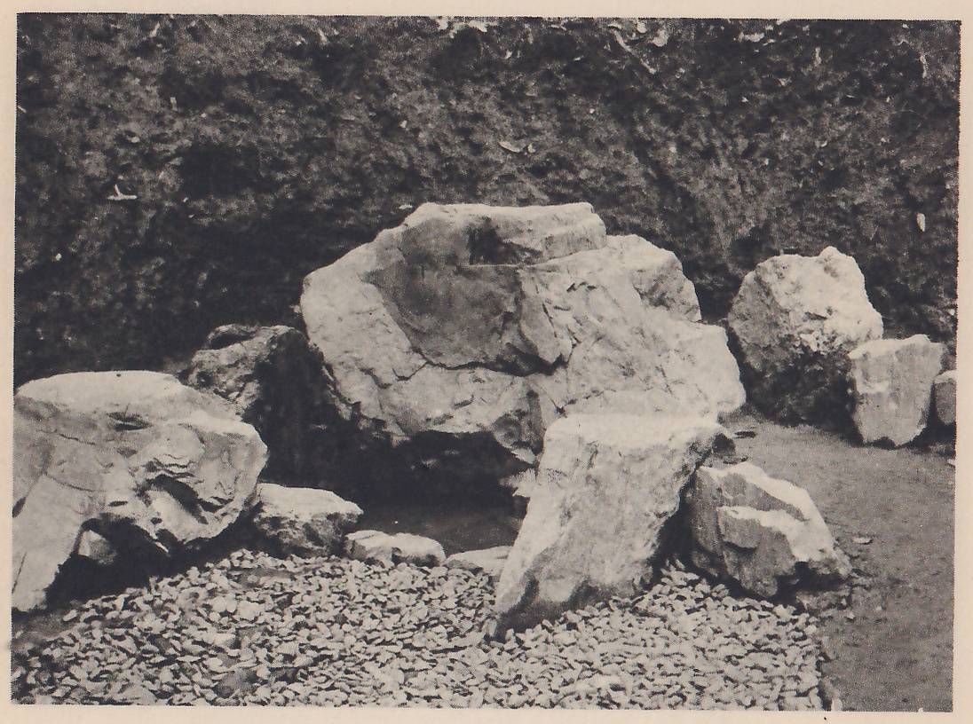 Photograph of a group of large, rough rocks standing on a bed of small pebbles in front of a bank of dark moss: a big central rock with smaller ones leaning against it and a flat-topped rock at the left, with a few rocks at the right.