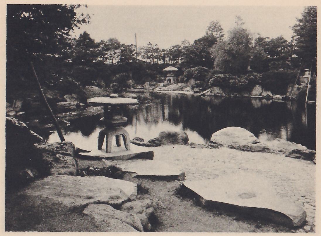 Photograph of a pond garden with a stone lantern with a wide round cap and curved legs standing on a flat rock at the water’s edge at the left of center, and a second lantern of the same kind small in the distance on the far shore. Large flat stones lie in the foreground, a leaning pole crosses the upper left, and trees surround the pond.