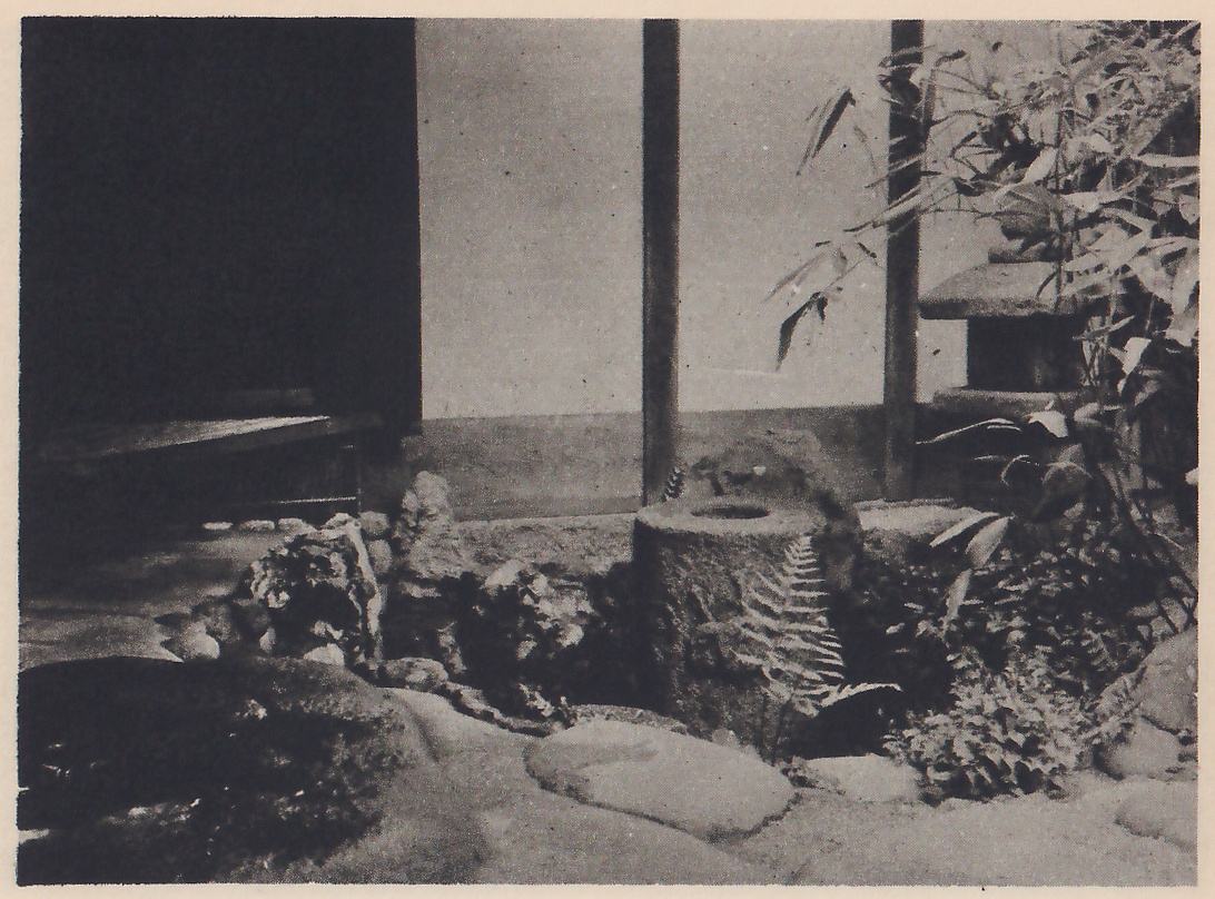Photograph of a short, rough stone post with a round hollow in its top, standing among rocks, a fern and low plants beside the verandah of a house; a flat stone lies in the foreground, a stone lantern shows at the right behind bamboo leaves, and the dark opening of a room is at the left.