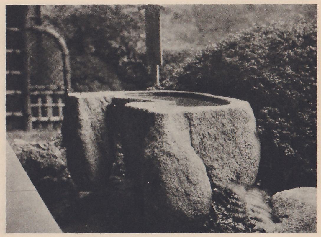 Photograph of a large rough stone basin holding water, with a second rough stone standing against its left side and a wooden spout above it, set among a rounded shrub, ferns and rocks. A lattice screen with an arched top stands at the left.