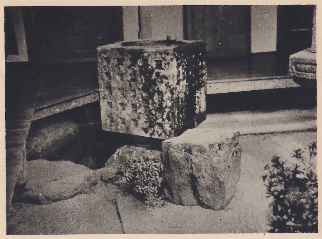 Photograph of a square stone basin with rows of small square holes in its sides, beside the raised floor of a house, with a rough upright stone in front of it; a flat stone lies at the left, a small shrub at the right, and smooth pale ground in front.