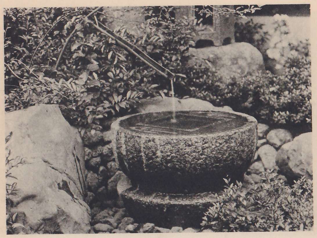 Photograph of a wide, shallow round stone basin full of water on a low round stone, with a thin stream of water falling into it from the end of a slanting bamboo pipe above. Large rocks, small round stones and leafy shrubs surround it, and part of a stone lantern shows at the upper right.