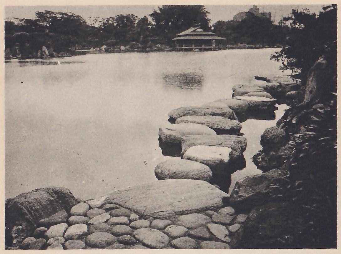 Photograph of a wide pond with a line of large rounded stepping-stones running out into the water at the right, a shore of small round stones and a large flat rock in the foreground, and a low roofed pavilion on the far shore among trees.