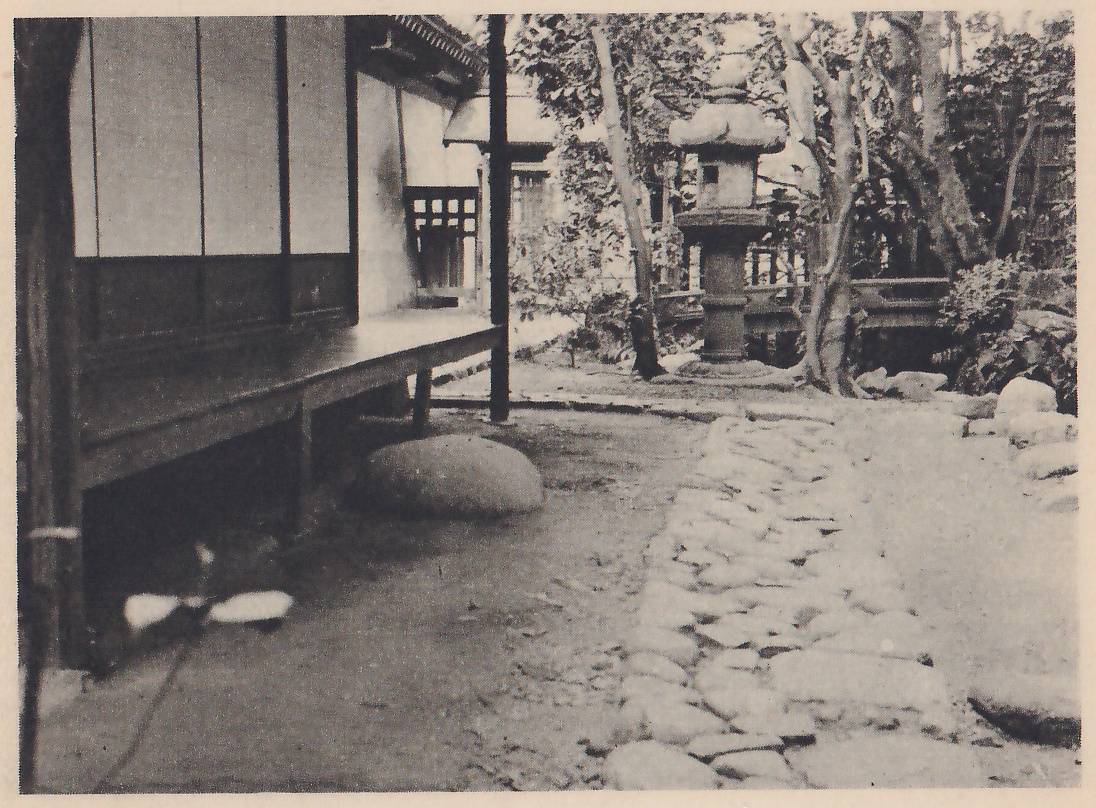 Photograph of a garden path of rounded cobbles laid in a broad band leading back along the raised wooden verandah of a building at the left, with sliding paper screens above it; a large rounded stone lies under the verandah, a tall stone lantern stands in the middle distance and trees and shrubs fill the background.