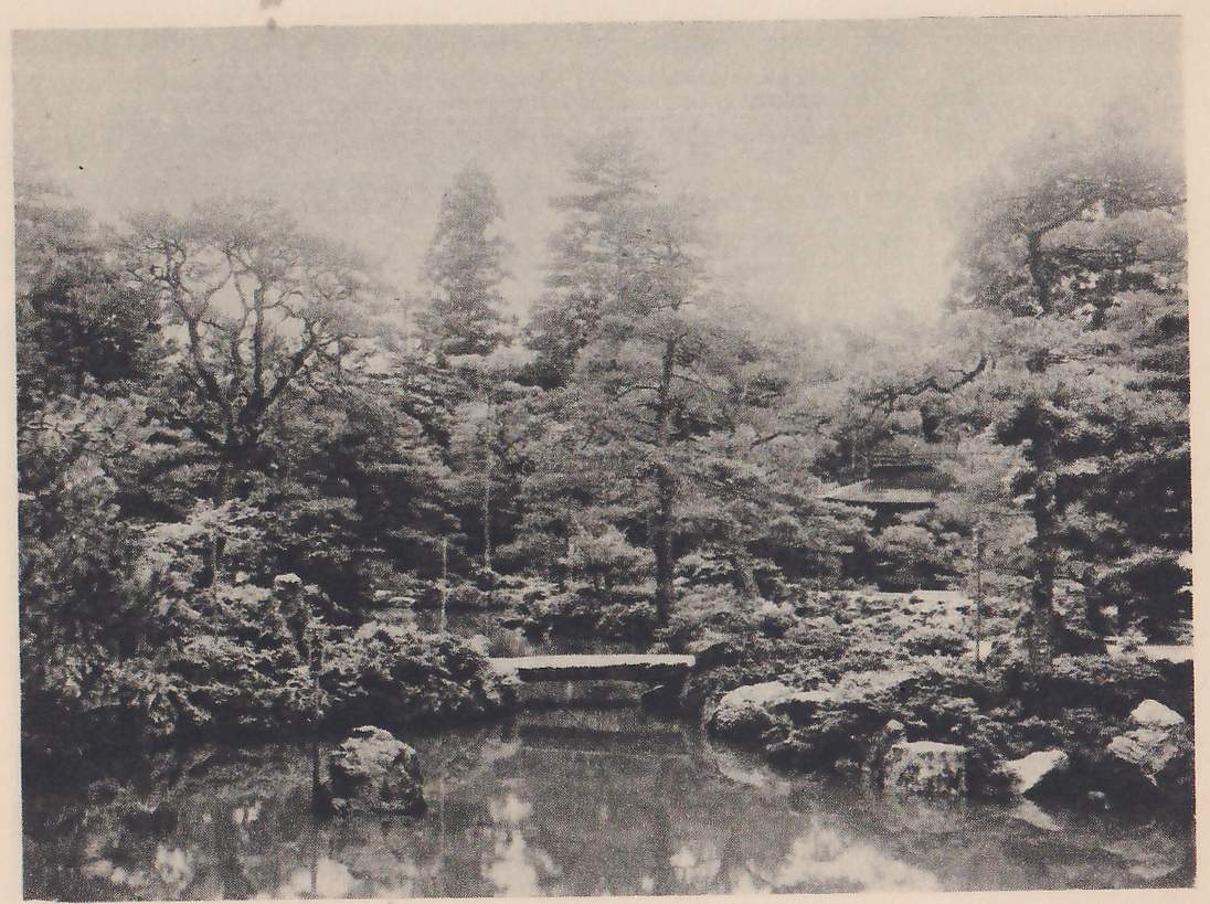 Photograph of a pond garden: a low flat stone bridge crosses a narrow gap at the back, with rocks and shrubs on both banks, tall pines and other trees behind, and the corner of a roof showing among them at the right.