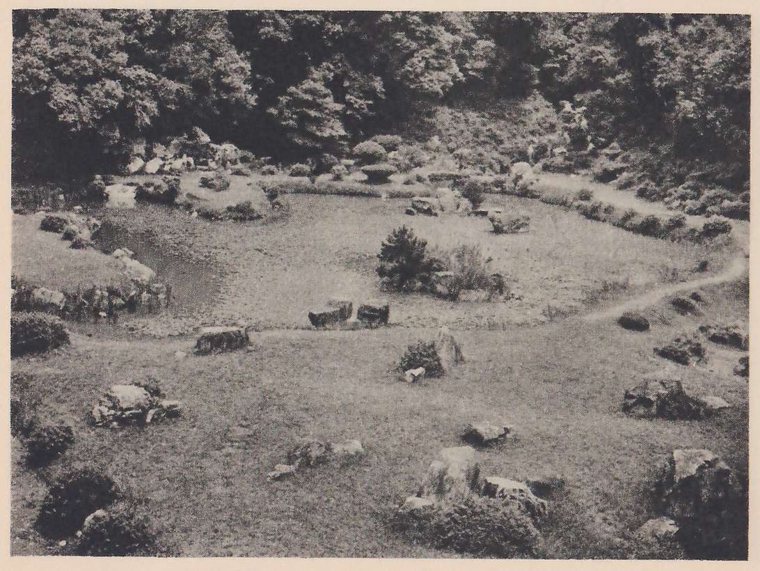 Photograph of a garden of open grassy ground with a strip of dark water at the left and a small clump of shrubs at the center; rocks are scattered across the grass, a worn path curves around the right side, and wooded hills rise behind.