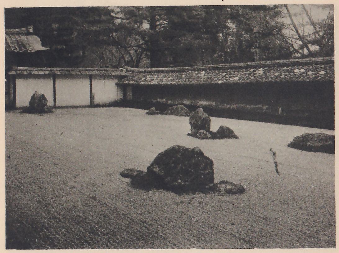 Photograph of a wide bed of raked gravel with groups of rocks standing in it, a large dark rock in the foreground and others farther back, enclosed by a long low wall with a tiled top, a roof corner at the left and trees behind.