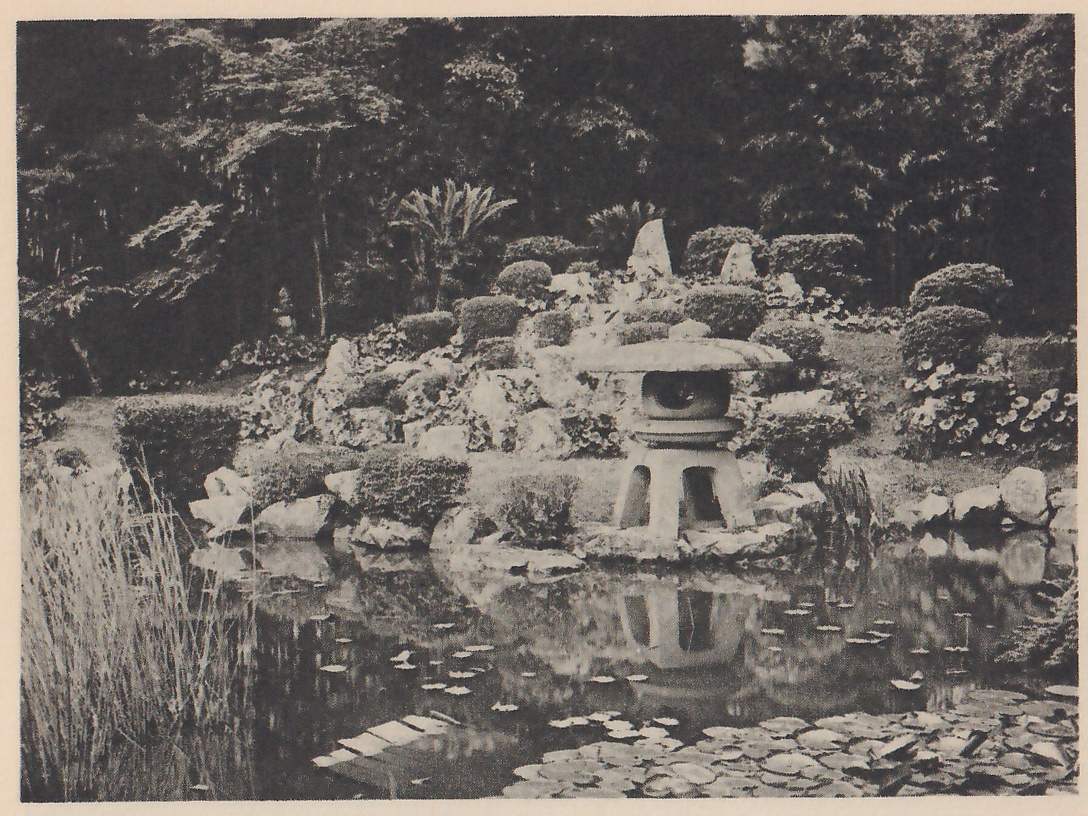 Photograph of a pond with water lilies and tall reeds at the left, and on the far bank a rocky mound planted with clipped shrubs and small palms, with a stone lantern with a wide round cap standing on the rocks at the water’s edge and reflected below; tall trees rise behind.