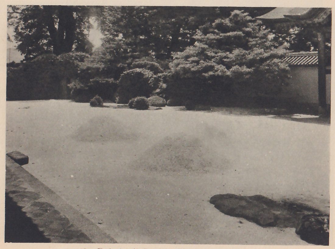 Photograph of a wide bed of white sand with two low raised mounds in it, a flat dark rock at the lower right and a stone edging along the left, enclosed at the back by clipped shrubs and a pine, with a plastered wall with a tiled top and the eave of a roof at the right.