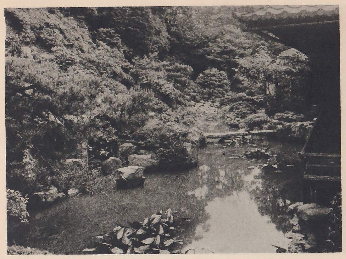 Photograph of a pond running back between a rocky, pine-covered slope at the left and the eave and dark side of a building at the upper right, with a low flat stone bridge across the far end, a flat rock in the water at the left, and broad-leaved water plants in the foreground.